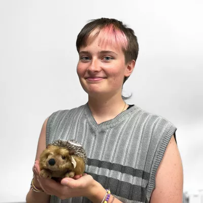 Une jeune personne avec une mèche de cheveux roses tient un hérisson en peluche dans ses bras, affichant un sourire joyeux.